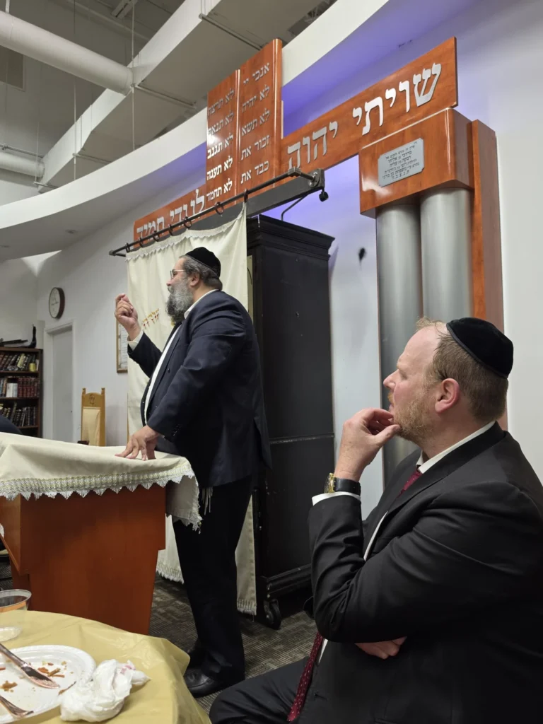 A rabbi speaking from the podium in front of the aron kodesh while a guest listens